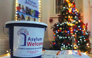 Image shows on an Asylum Welcome donation bucket on a table with Christmas cards in front of a Christmas tree.