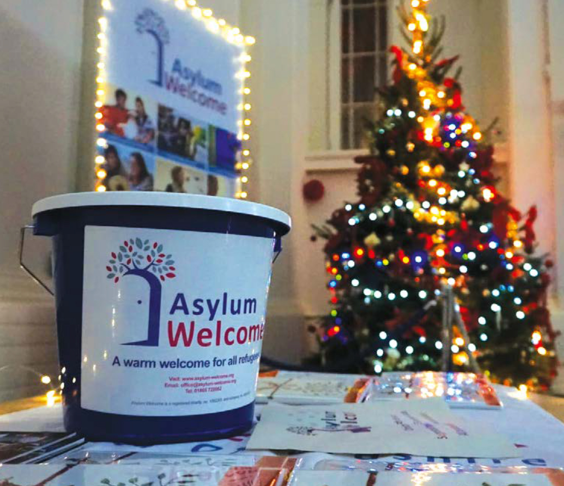 Image shows on an Asylum Welcome donation bucket on a table with Christmas cards in front of a Christmas tree.