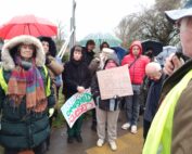 A group of protesters stand closely together in wet weather, listening to a speaker in a high-visibility vest, with several people holding handmade “Keep Campsfield Closed” signs and umbrellas in the background.