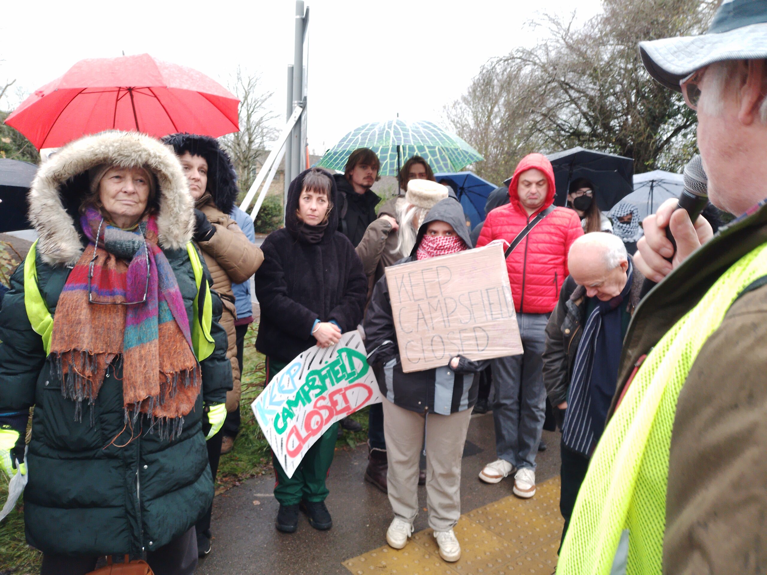 A group of protesters stand closely together in wet weather, listening to a speaker in a high-visibility vest, with several people holding handmade “Keep Campsfield Closed” signs and umbrellas in the background.